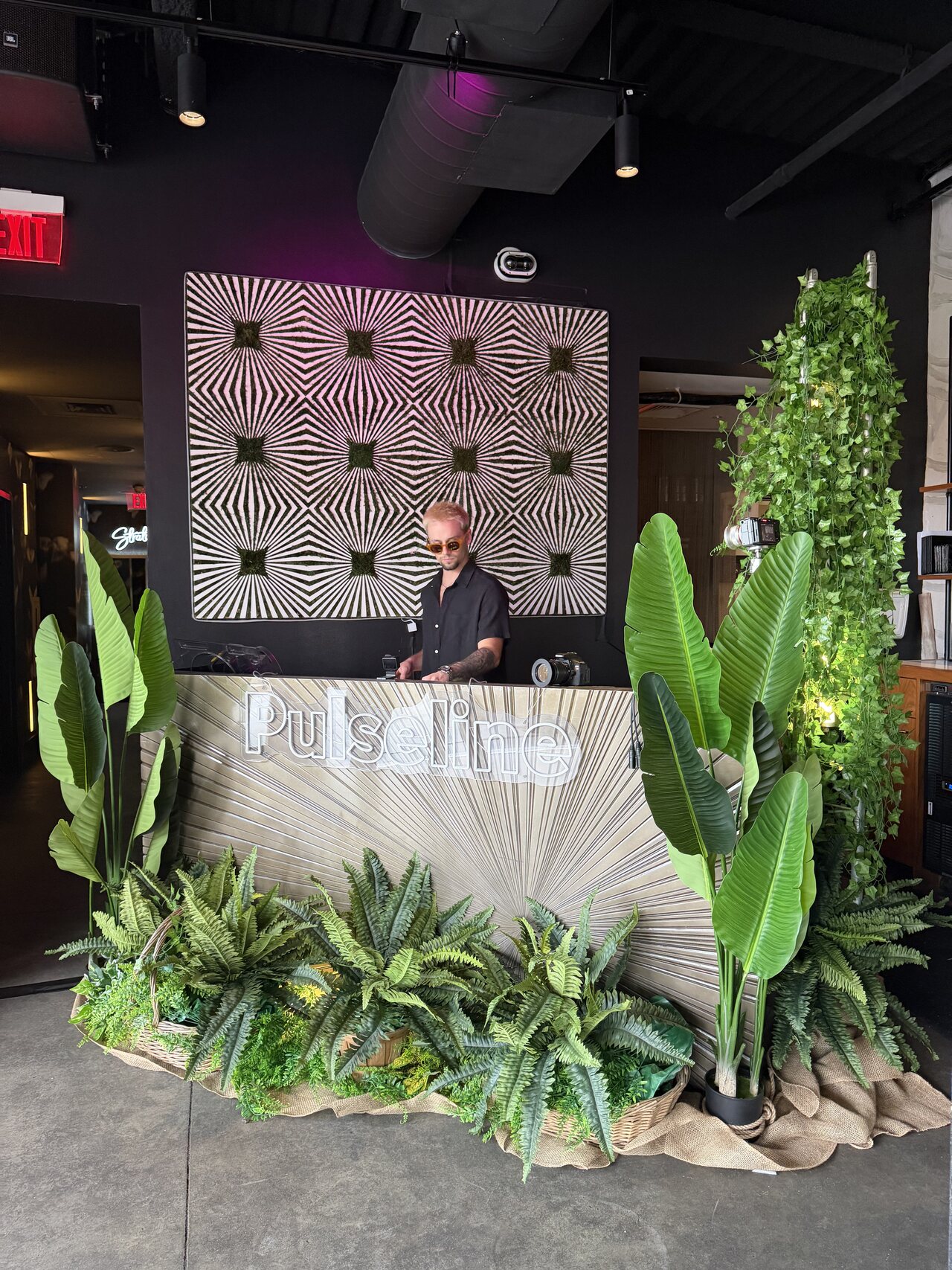 A DJ booth dressed in plants and a custom Pulseline facade at a daytime event