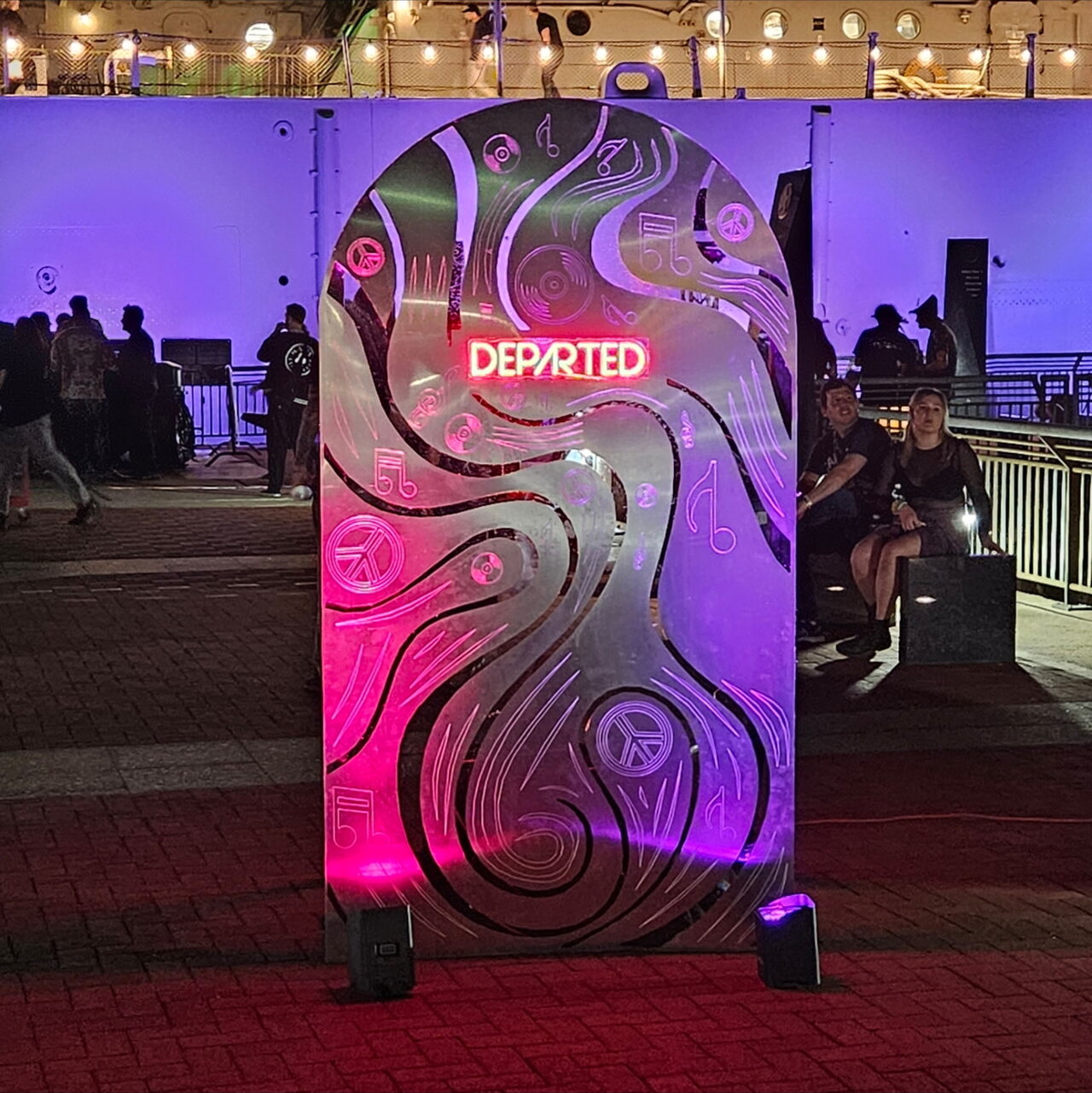 A custom-fabricated metal sign with a neon Departed logo, lit in pink and purple